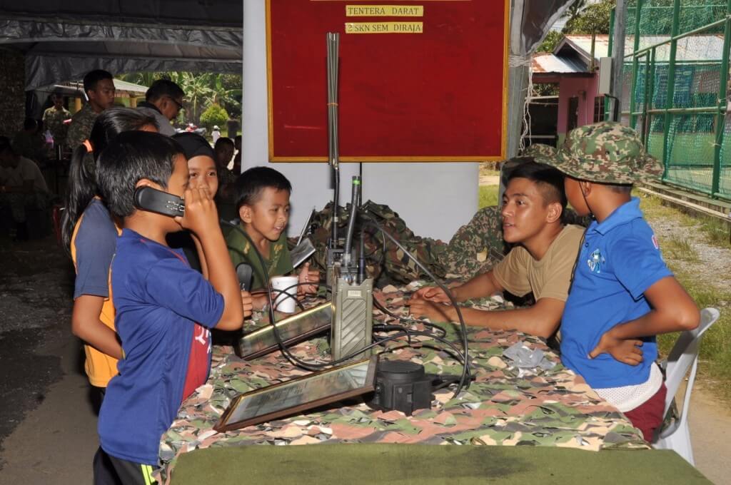 Kanak-kanak lelaki ini berlakon menggunakan peralatan komunikasi tentera sambil diperhatikan oleh anggota tentera yang bertugas